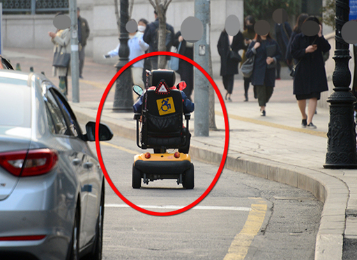 전동휠체어를 탄 한 시민이이 차도에서 저속으로 오르고 있다. 전동 휠체어는 인도로만 다녀야 한다.