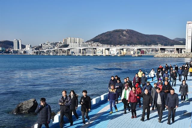 부산 영도구 흰여울길을 찾은 관광객들이 시원한 바닷바람을 맞으며 무리를 지어 길을 걷고 있다. 관광객 뒤로 탁 트인 바다와 산 주변에 옹기종기 모여 있는 집들이 정겨운 조화를 이룬다.