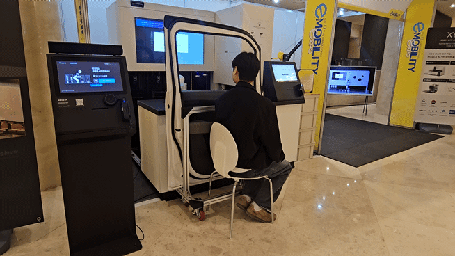 시연에 나선 직원이 25일 제주 신화월드에서 열린 국제 e-모빌리티 엑스포 부스에서 의자에 앉아 창문 너머로 음료를 받고 있다. [사진=설재윤 기자]